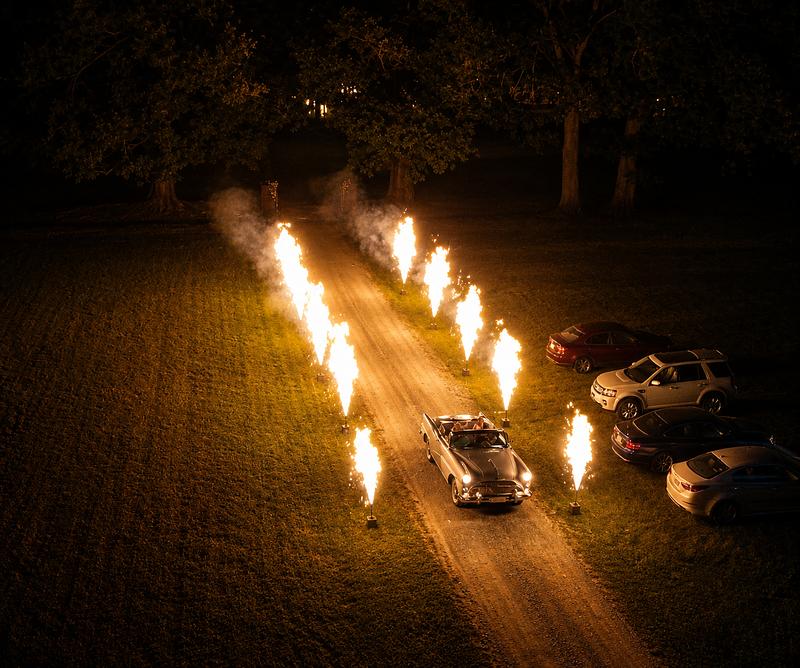 Classic convertible driving between gerb flame columns lining a wedding driveway at night